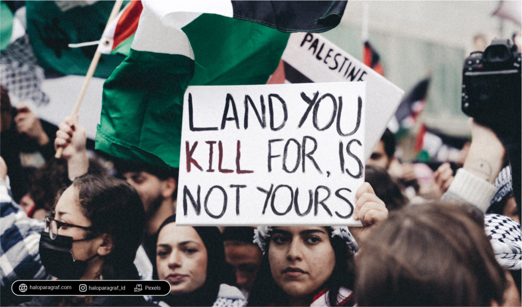 Potret kerumunan masa; salah satu orang di antaranya memegang papan bertuliskan "LAND YOU KILL FOR, IS NOT YOURS"